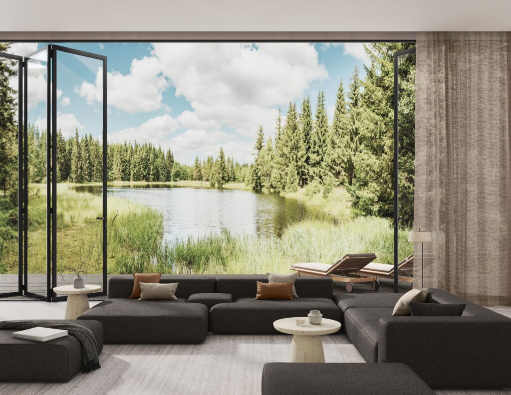 A modern living room with a dark grey modular sectional sofa opens onto a terrace via black framed folding glass doors overlooking a lake surrounded by pine forest Two wooden lounge chairs sit on the deck
